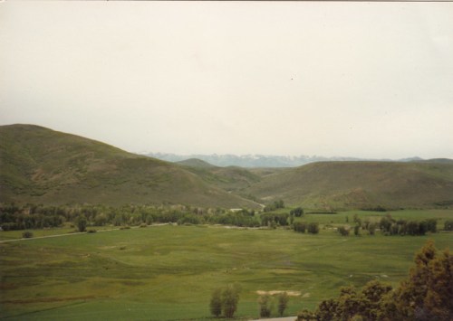 A serene landscape featuring rolling green hills, a river, and distant mountains under a cloudy sky.