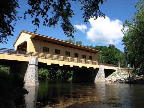 A wooden covered bridge spans a river, surrounded by greenery and under a blue sky with scattered clouds.