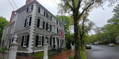 Historic gray house with black shutters and a flag, surrounded by trees on a quiet street on a cloudy day.