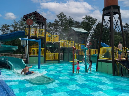 A vibrant water park scene with slides, a splash pad, and children playing in a blue pool under a sunny sky.