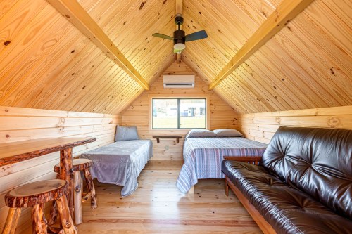 Cozy wooden interior of a cabin with two beds, a sofa, and a small table under a sloped ceiling.