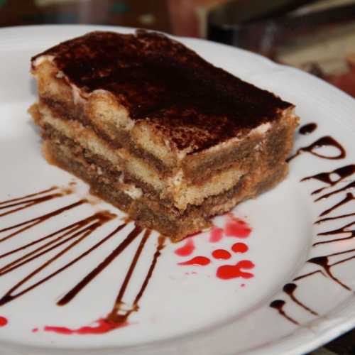 A slice of tiramisu on a white plate, drizzled with chocolate sauce and decorated with red splatters.