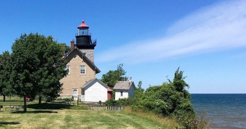 A lighthouse beside a calm lake, surrounded by green trees and a clear blue sky.