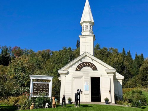 A small white chapel with a steeple, surrounded by trees, featuring dog statues and a welcoming sign.
