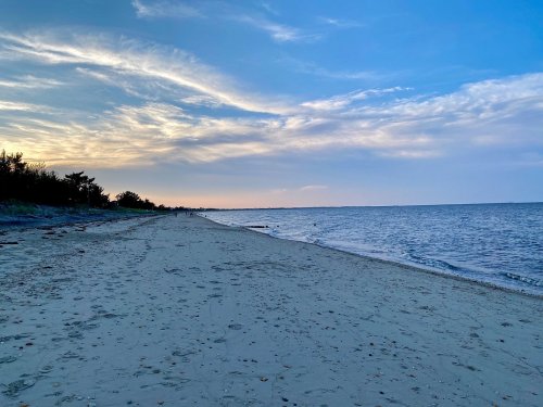 A serene beach at sunset, with soft sand, gentle waves, and a clear sky filled with wispy clouds.