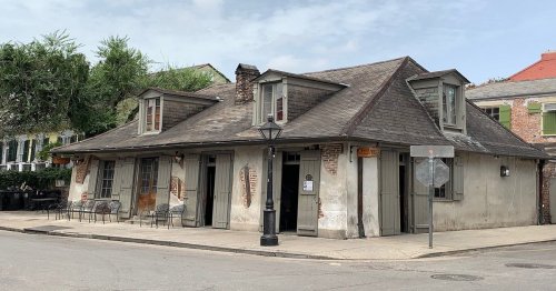 Historic building with a sloped roof, wooden doors, and outdoor seating, set on a quiet street with greenery.
