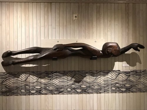 A wooden sculpture of a swimmer, elongated and stylized, displayed against a wooden wall with a wave pattern below.