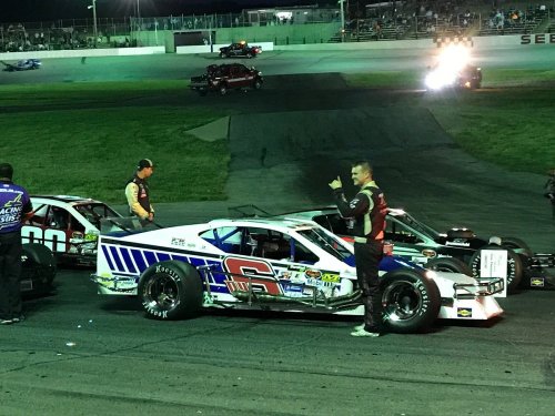 A night scene at a racetrack with cars lined up and drivers interacting, illuminated by track lights.