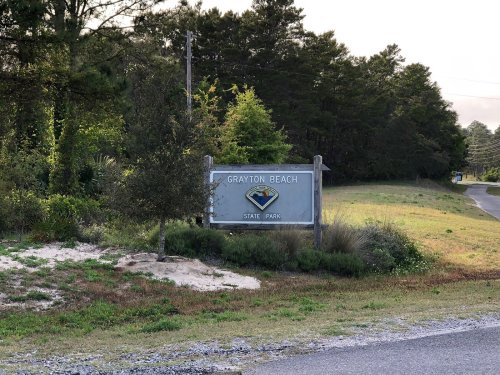 Sign for Grayton Beach State Park, surrounded by greenery and a gravel road in the background.