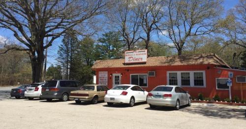 A red diner building with a sign, surrounded by trees and parked cars in a sunny outdoor setting.