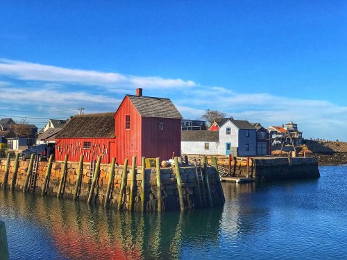 A red building by the water, reflecting in the calm sea, with white houses and a blue sky in the background.