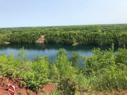 A serene lake surrounded by lush green trees under a clear blue sky. A red sign is visible in the foreground.