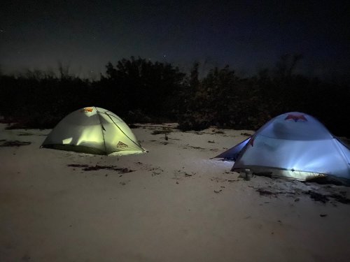 Two tents illuminated at night, set against a dark, wooded background with a snowy ground.