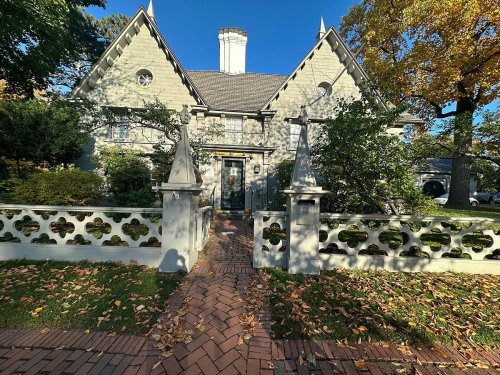 A large, gray house with a steep roof, surrounded by trees and a decorative fence, on a sunny autumn day.