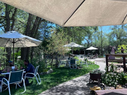 A sunny outdoor café with tables under umbrellas, surrounded by greenery and trees. People are enjoying their time.