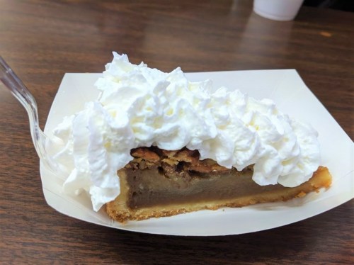 A slice of pie topped with whipped cream, featuring a nutty filling and a golden crust, served on a paper plate.