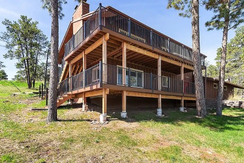 A wooden house with multiple decks, elevated on pillars, surrounded by trees and grassy land.