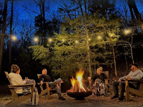A group of four people sitting around a campfire at night, surrounded by trees and string lights.