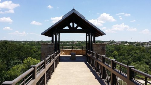 A wooden pavilion with a peaked roof overlooks a lush green landscape under a blue sky with scattered clouds.