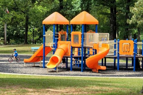 Colorful playground with orange slides and climbing structures, surrounded by green trees and a child riding a bike nearby.