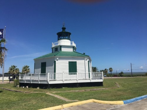 A white lighthouse with a green roof, surrounded by grass and palm trees, under a clear blue sky.