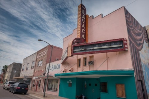 A vintage theater with a marquee, pink exterior, and nearby buildings in a small town under a cloudy sky.