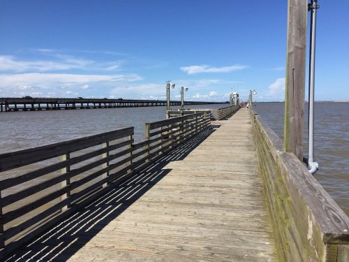 A wooden pier extends over calm water under a clear blue sky, with shadows cast by the railing.