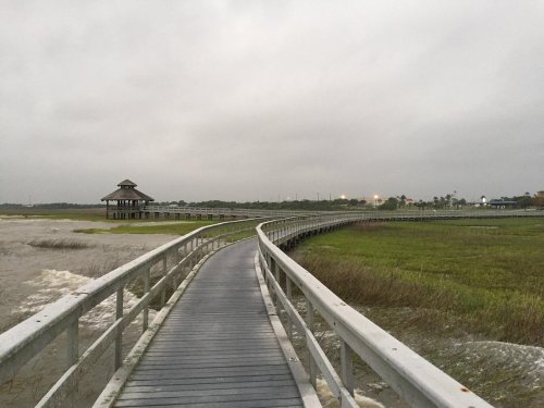 A winding boardwalk leads to a gazebo over a marshy landscape under a cloudy sky.