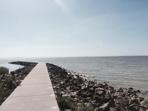 A concrete walkway extends over rocky shorelines towards a calm sea under a clear blue sky.