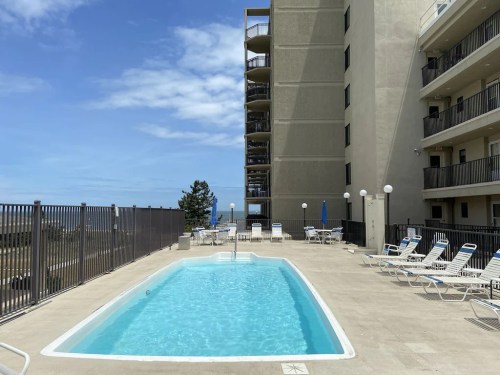 A clear swimming pool surrounded by lounge chairs, with a tall building and blue sky in the background.