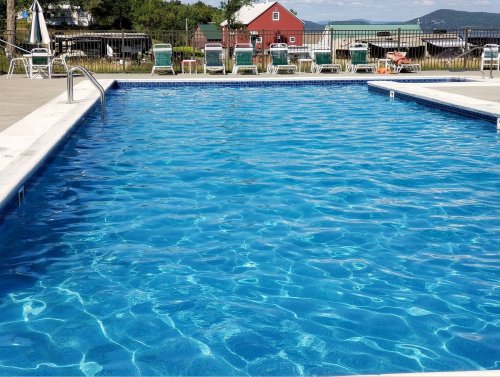 A clear blue swimming pool with lounge chairs around it, set against a backdrop of green hills and a red building.