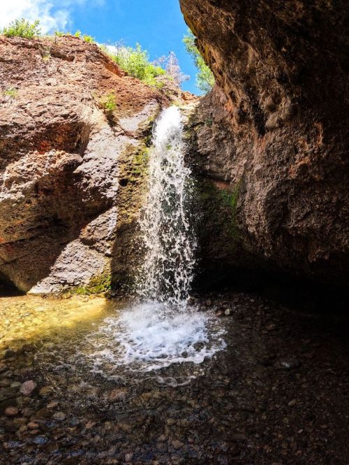 A small waterfall cascades over rocky terrain into a clear pool below, surrounded by lush greenery and blue sky.