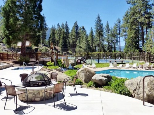 A sunny outdoor pool area surrounded by trees, featuring a fire pit and lounge chairs.