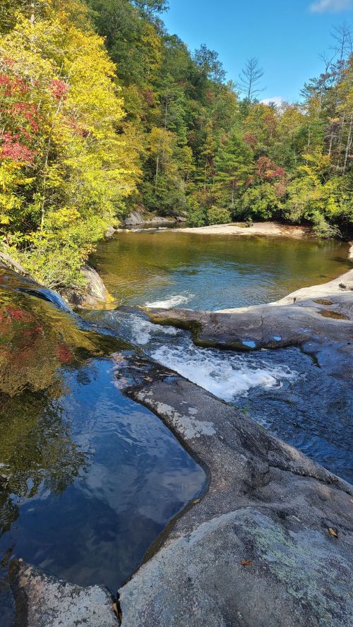 A serene river flows over rocks, surrounded by vibrant autumn foliage and a clear blue sky.