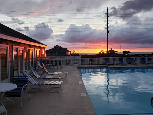Sunset reflecting on a poolside with lounge chairs, surrounded by buildings and a cloudy sky.