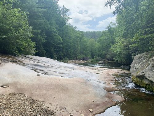A serene river scene with smooth rocks, surrounded by lush green trees under a partly cloudy sky.