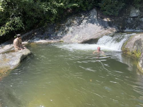 Two people enjoy a natural swimming hole with a small waterfall surrounded by rocks and greenery.