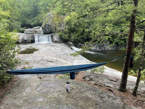 A hammock is set up between two trees near a rocky area, with a waterfall and a water bottle in the foreground.