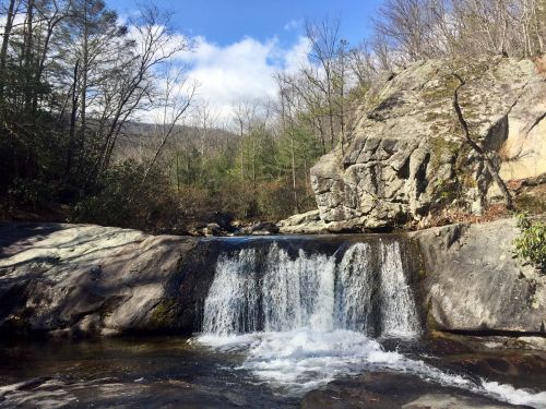 A serene waterfall cascades over rocks, surrounded by trees and a clear blue sky in a tranquil natural setting.