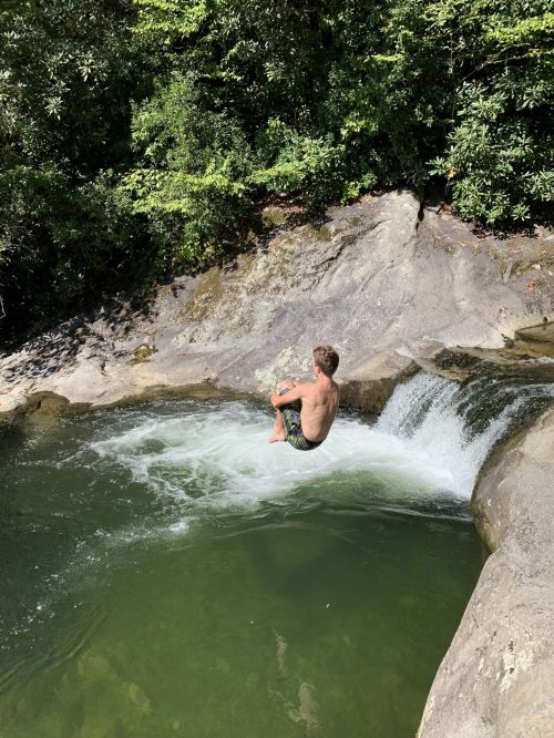 A person sits on the edge of a rocky ledge above a green pool, preparing to jump into the water below.