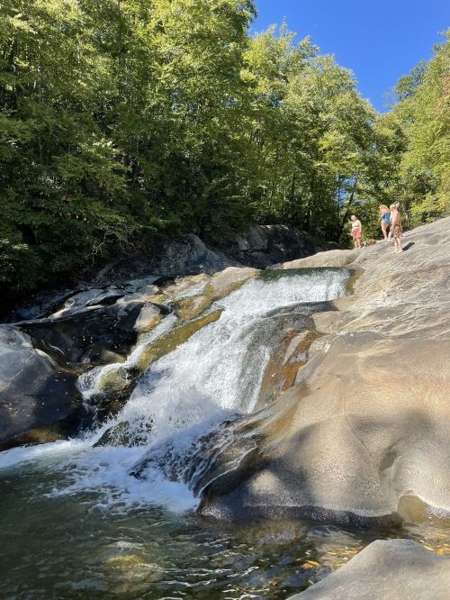 A serene waterfall cascades over smooth rocks, with people enjoying the natural scenery in a lush green setting.