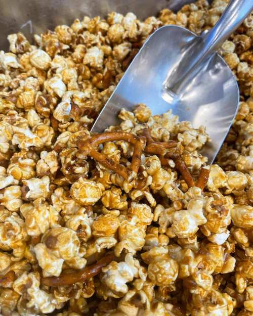 A close-up of caramel popcorn mixed with pretzels, with a metal scoop resting on top.