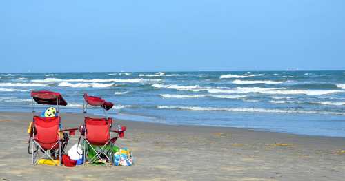 Two red beach chairs facing the ocean, with a soccer ball and beach toys nearby on a sandy shore.