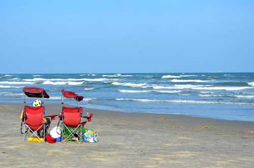 Two red beach chairs facing the ocean, with gentle waves and a clear blue sky in the background.