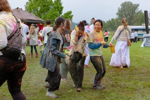 A group of people covered in colorful powder celebrate joyfully at an outdoor festival, surrounded by greenery.