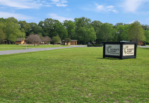 A grassy area with a sign for Poverty Point and several buildings in the background under a clear blue sky.