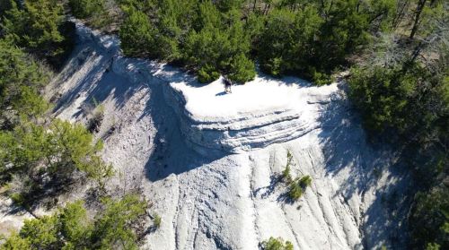 Aerial view of a rocky cliff surrounded by green trees, with a person standing on the edge of the white terrain.