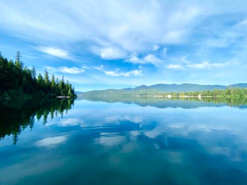 A serene lake reflecting blue skies and clouds, surrounded by lush green trees and distant mountains.