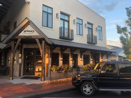 A modern building with a sign reading "primary" and a black SUV parked in front, surrounded by flowers.