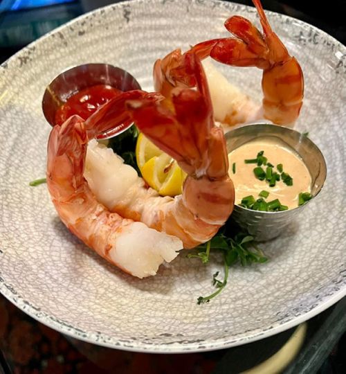 A plate with two large shrimp, lemon slices, and two dipping sauces, garnished with greens.